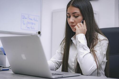 A photo of a woman sitting at a desk, looking at a laptop, and holding a cell phone to her ear.